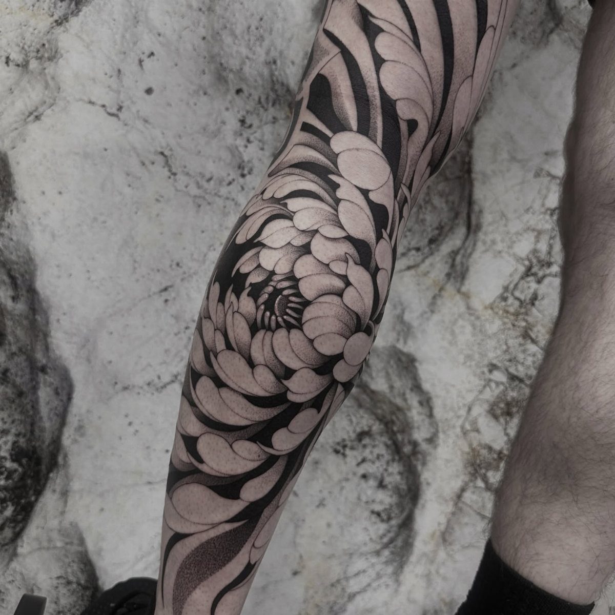 17 Flower Tattoo Ideas That Work in Every Style 9 A large black and grey chrysanthemum tattoo covering a person's leg against marble.