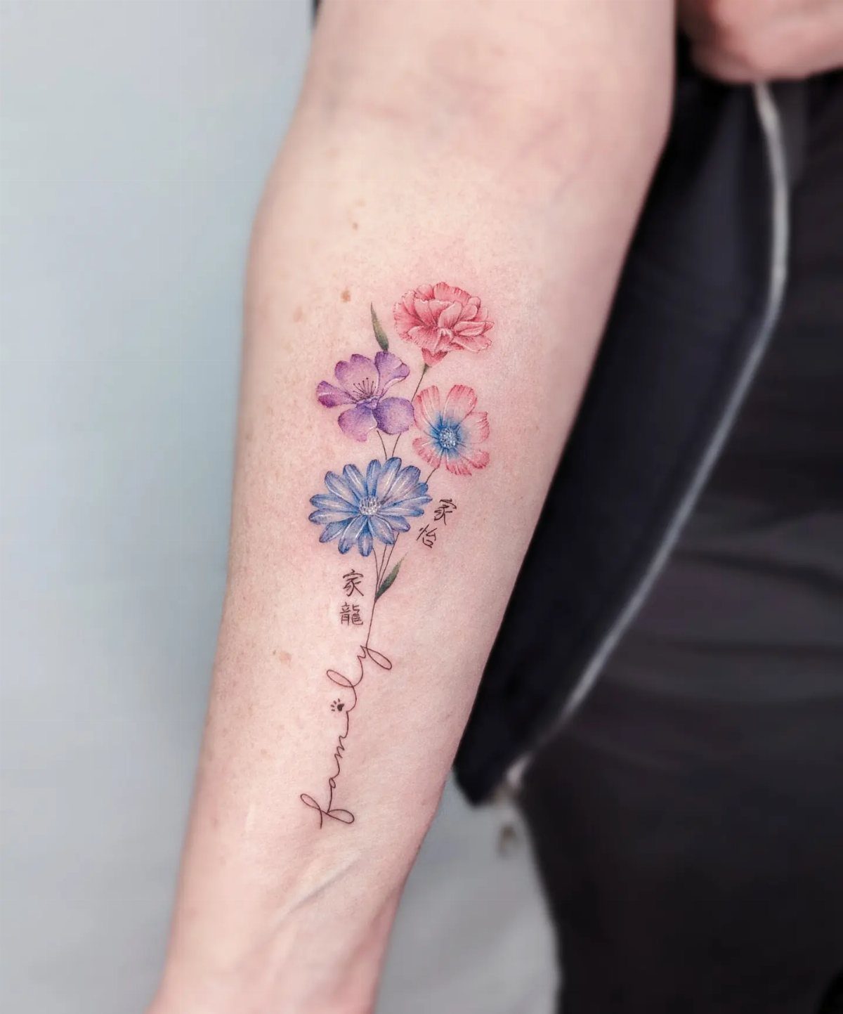 17 Flower Tattoo Ideas That Work in Every Style 16 A colorful floral tattoo on a forearm with delicate stems turning into cursive text.