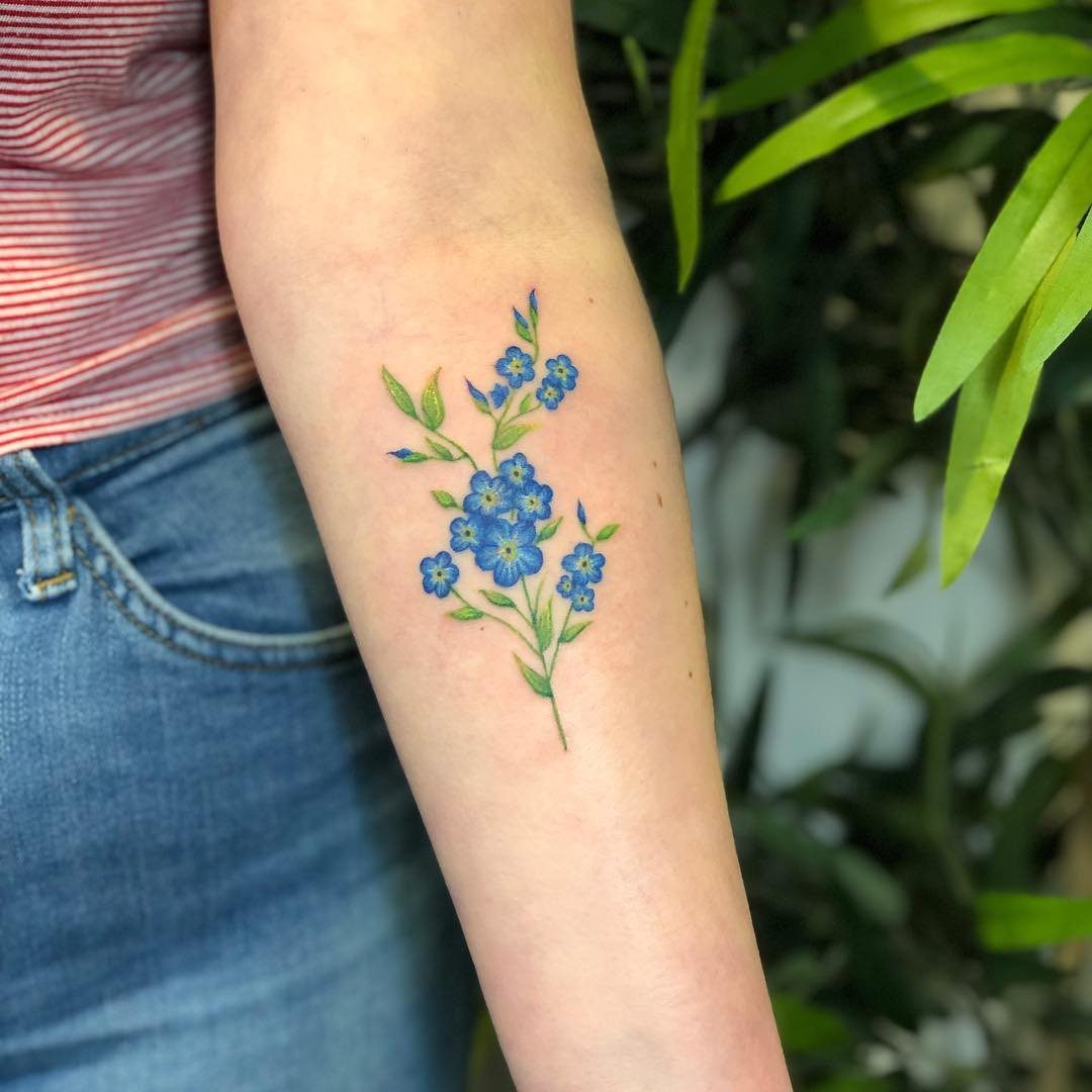 17 Flower Tattoo Ideas That Work in Every Style 11 A blue forget-me-not floral tattoo on a person's forearm against a green leafy background.