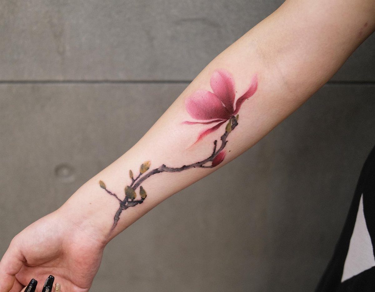 17 Flower Tattoo Ideas That Work in Every Style 8 A pink magnolia flower tattoo with a dark branch stretching across a person's forearm.