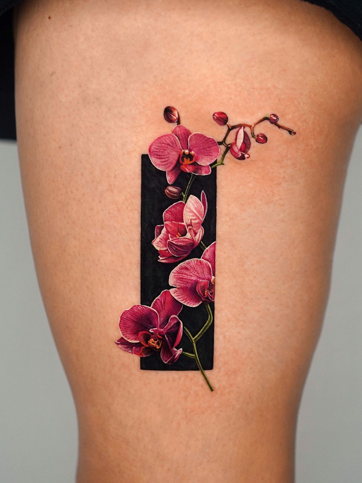17 Flower Tattoo Ideas That Work in Every Style 6 A realistic pink orchid tattoo with dark buds set against a black rectangular background on skin.