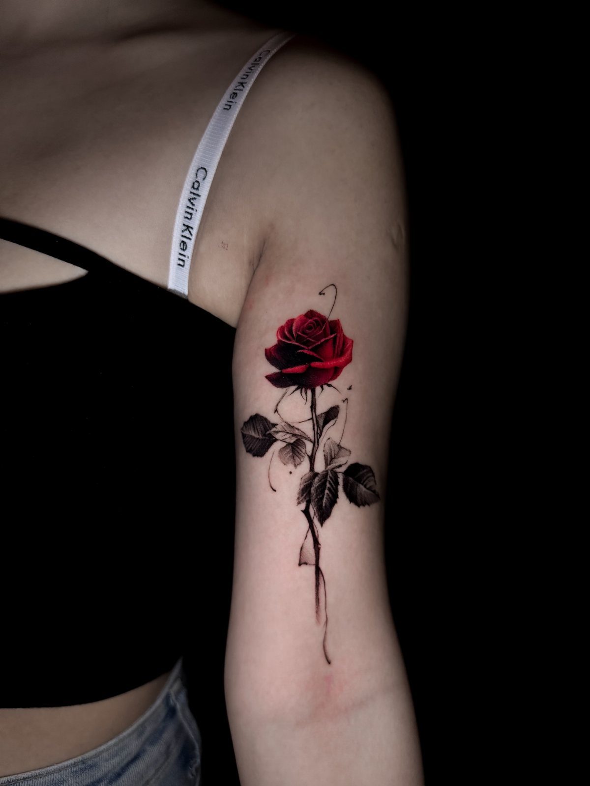 17 Flower Tattoo Ideas That Work in Every Style 3 A vibrant red rose tattoo with black leaves and stem on a person's upper arm.