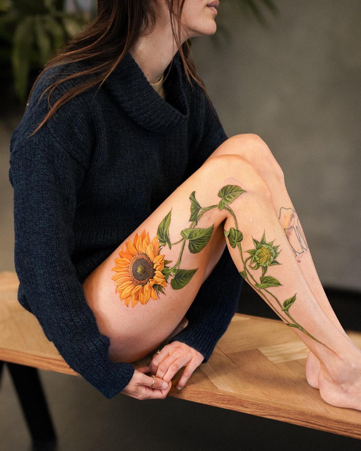 17 Flower Tattoo Ideas That Work in Every Style 4 A large, vibrant sunflower tattoo with green leaves winding down a person's leg.